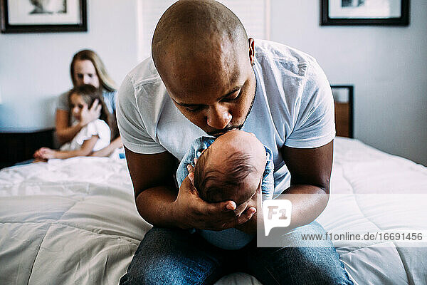 dad kissing newborn on bed with mom and daughter in back ground