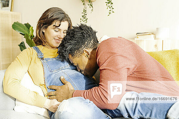 Ehemann küsst seine schwangere Frau auf einem Sofa. Interracial Paar Konzept