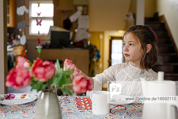 A small child in a lace dress sits alone at a table set for tea party