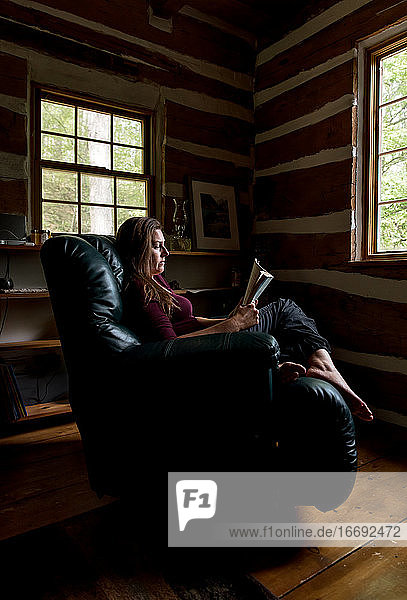 Frau liest im Ledersessel in einer rustikalen Blockhütte.