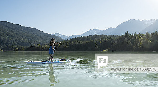 Barfüßige Frau auf einem Paddelbrett auf einem See vor einem grünen Bergkamm an einem sonnigen Sommertag in British Columbia  Kanada