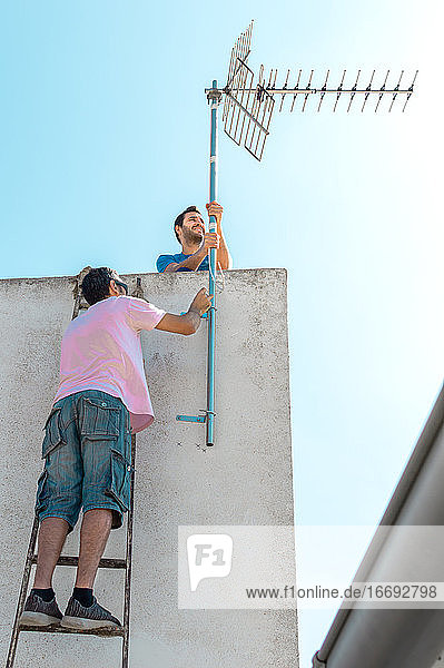 Zwei Männer installieren an einem sonnigen Tag eine Fernsehantenne auf dem Hochdach eines Landhauses.
