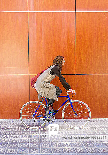 Hübsche junge Frau mit Brille fährt mit dem Fahrrad gegen eine rote Wand.
