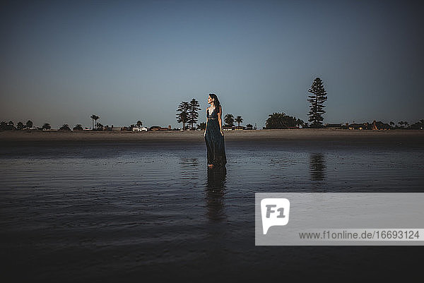 Schöne Frau in langem Kleid steht am Strand bei Sonnenuntergang