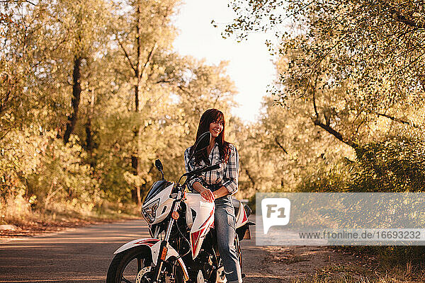 Smiling young woman sitting on motorcycle on country road amidst trees
