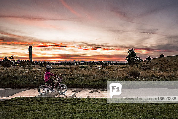 young girl rides a bike along the sidewalk in a park at sunset
