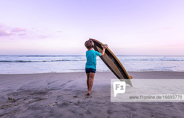 Rückansicht 4 Jahre altes Kind trägt ein hölzernes Surfbrett am Strand