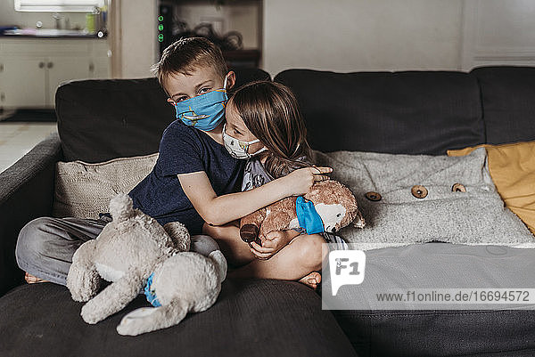 Junges Mädchen und Junge im Schulalter mit Masken  die sich auf der Couch umarmen und lächeln