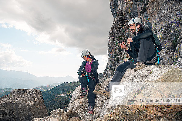 Konzept: Abenteuer. Ein Paar Kletterer mit Helm und Gurtzeug. Sie ruhen sich auf einem Felsen sitzend aus. Sie benutzen das Smartphone und schauen auf den Horizont. Via ferrata in den Bergen.