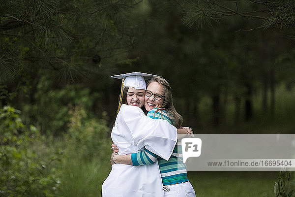 Glückliche Absolventin mit weißer Kappe und Kittel umarmt fest ihre Mutter