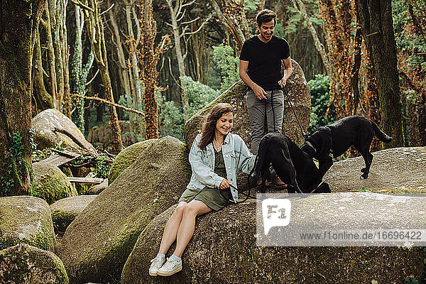 Young couple with two dogs having fun in forest while sitting on rock