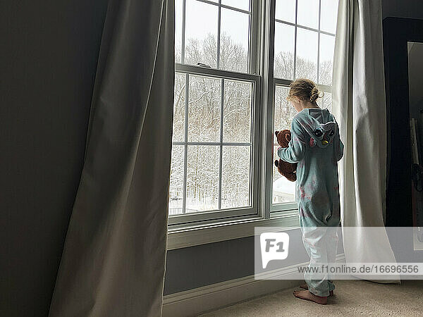 Rear View of Girl in Pajama Onesie Holding Teddy Looking Out Window