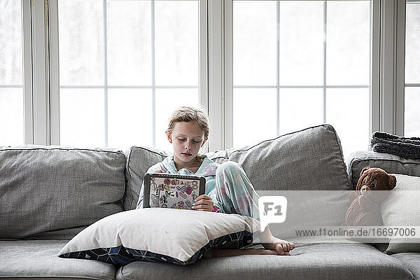 Blonde Girl Home Sick From School Plays Tablet on Couch With Teddy