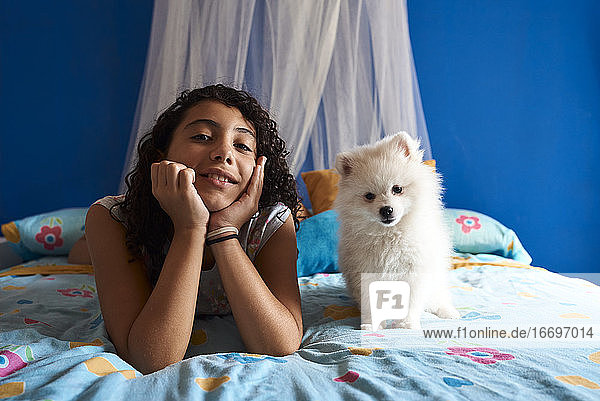 ein kleines Mädchen und ihr weißer Hund sitzen auf dem Bett und schauen in die Kamera