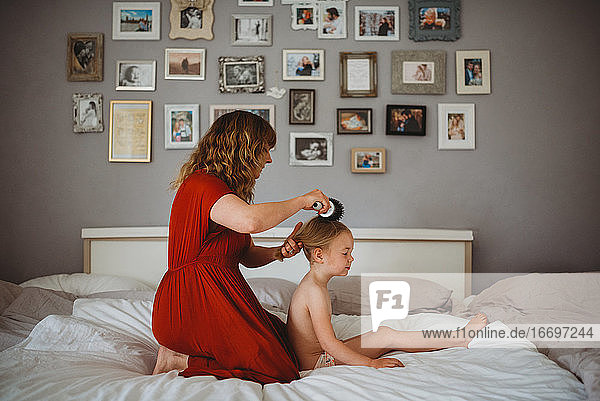 Mother brushing daughter's hair in family bed with photos on wall
