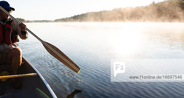 Ein Mann paddelt morgens mit seinem Kanu auf einem nebligen See in Ontario  Kanada.
