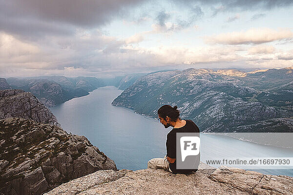 Mann schaut neugierig nach unten  sitzt in einem Felsen am Rande einer Klippe am Preikestolen  Norwegen