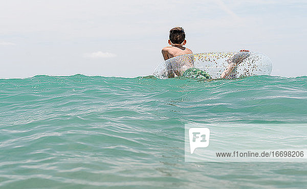 entspannter weißer Junge schwimmt in einem Schwimmer im Meer und genießt die ruhigen Wellen im Sommer. Rückwärts horizontales Foto