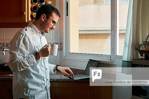 Junger Mann schaut auf seinen Laptop  während er in seiner Küche Kaffee trinkt