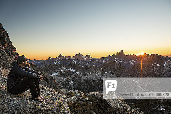 Aktiver  fitter Mann ruht sich auf einem Felsen in den Bergen aus und beobachtet den Sonnenuntergang.