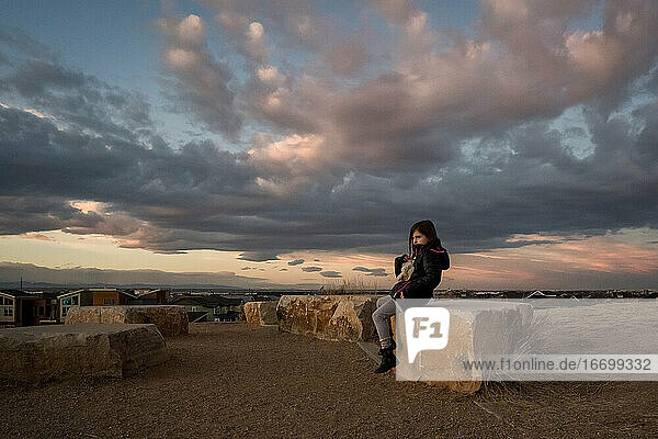 young girl sits on a rock at the top of a hill at sunset