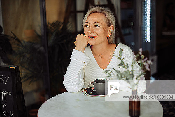 Blonde Frau lächelt  während sie mit einer Tasse Kaffee am Tisch im Café sitzt