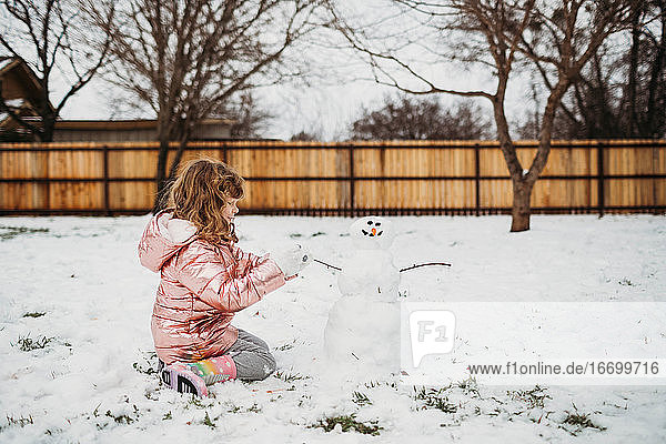 Blondes  gelocktes Mädchen sitzt draußen im Schnee und baut einen Schneemann