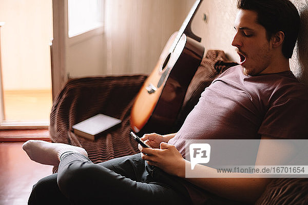 Junger Mann gähnt  während er sein Smartphone benutzt und zu Hause sitzt