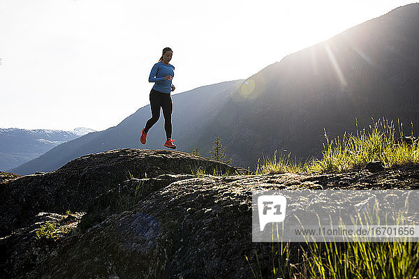 Ganzkörper-Sportlerin in Sportkleidung läuft auf Felsen in der Nähe von Bergen an einem sonnigen Abend auf dem Lande