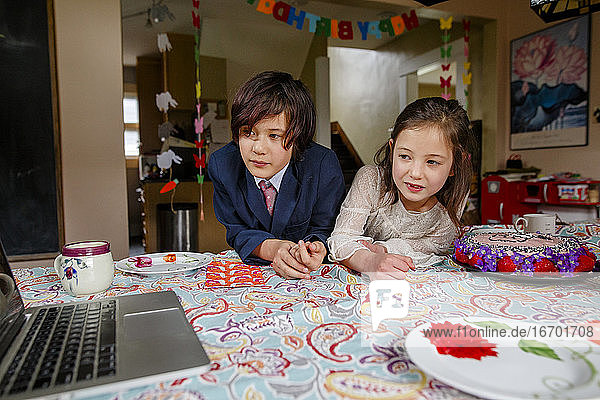 A boy and girl sit at table with birthday cake watching a computer