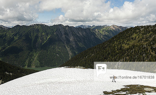 Von oben Seitenansicht von anonymen Forscher mit Rucksack zu Fuß auf schneebedeckten Hügeln gegen majestätischen bewaldeten Bergen in bewölkten Tag