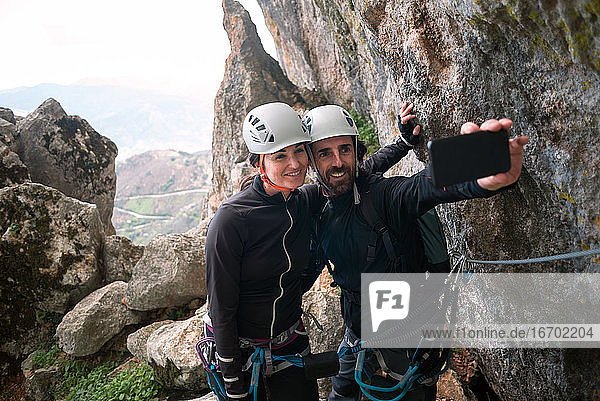 Concept: adventure. Pair of climbers with helmet and harness. Taking a selfie with the smart phone. At the top of the mountain. Via ferrata in rock.