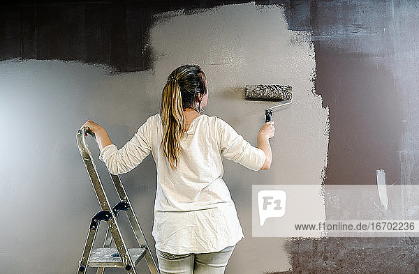 Frau klettert auf eine Leiter und fängt eine Farbrolle voller grauer Farbe auf einer braunen Wand. Der Maler fährt die Rolle hoch und runter und bedeckt die Wand mit grauer Farbe  die noch feucht ist. Horizonta