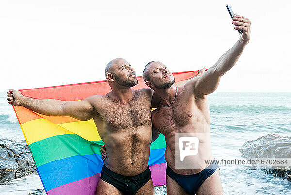 Lgbt pride concept. Gay bear couple holding rainbow flag with open arms taking a selfie photo.