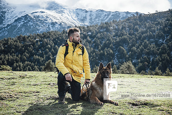 Young man with yellow jacket and backpack plays with German Shepherd dog in the mountains.