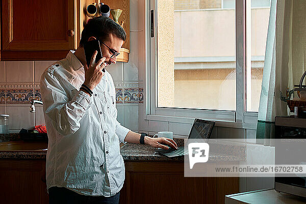 Junger Mann telefoniert mit seinem Smartphone und schaut auf seinen Laptop in der Küche