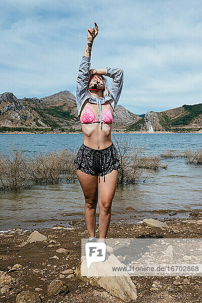 Junge Frau am Ufer eines Sees in Spanien in einem Bikini und Shorts. Stehend in einer entspannten Position mit ihren Armen oben