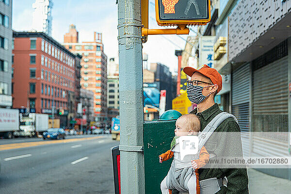 Father in face mask carrying baby girl while standing on sidewalk in city during COVID-19 outbreak