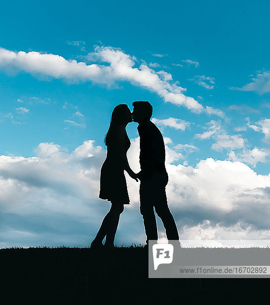 Silhouette of man and woman kissing against a blue sky with clouds.
