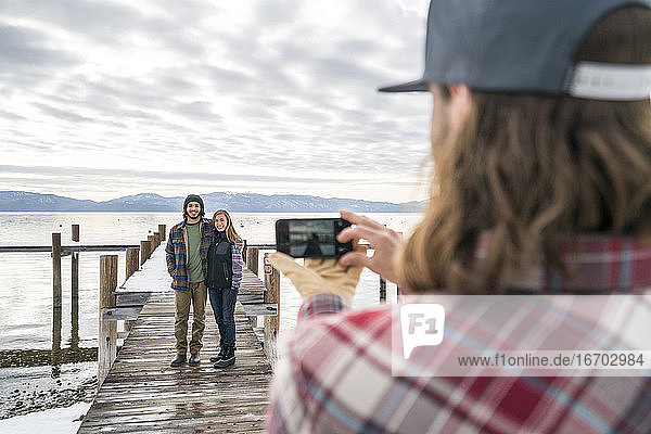 Ein Freund macht ein Handyfoto für ein Paar  das am Ende eines Piers steht