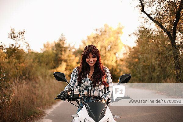 Porträt einer glücklichen jungen Frau  die auf einem Motorrad auf einer Landstraße sitzt
