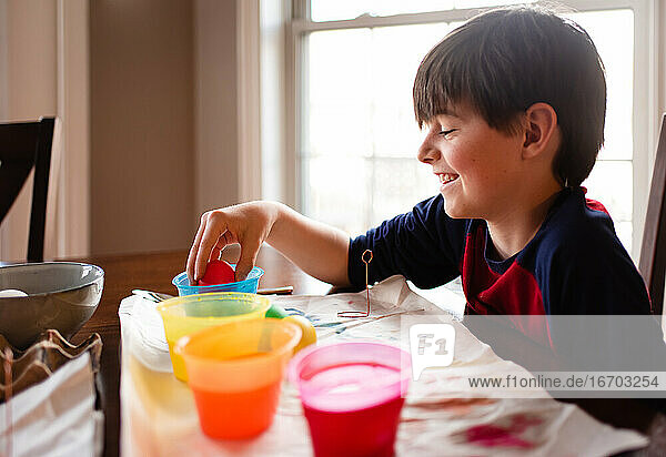Ein fröhlicher Junge taucht ein Ei in eine Schale mit Farbstoff  um es für Ostern zu färben.
