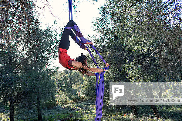 Junges Mädchen  Akrobatin. Übt Seiltanz in der Luft. Frau macht Zirkuskunststücke mit Kleidung im Wald. Skorpion-Stellung.