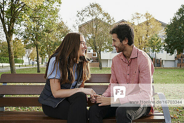 Junges Paar  das sich lächelnd auf einer Bank im Park sitzt und sich gegenseitig anstarrt. Romantisches Konzept