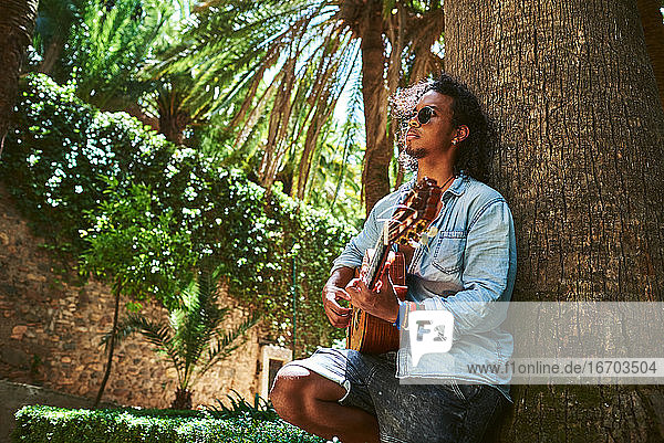 Junger Musiker mit klassischer Gitarre spielt im Schatten einiger Bäume in einem Park.