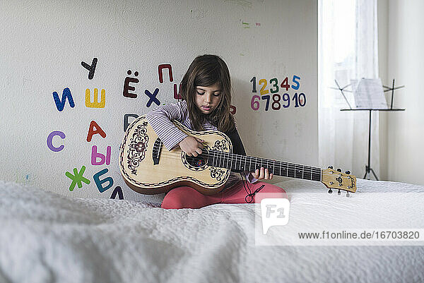 Junges Mädchen auf dem Bett spielt Gitarre vor einer Wand mit Alphabet-Aufklebern