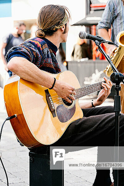 Mann mit zusammengebundenen langen Haaren singt und spielt Gitarre auf der Straße