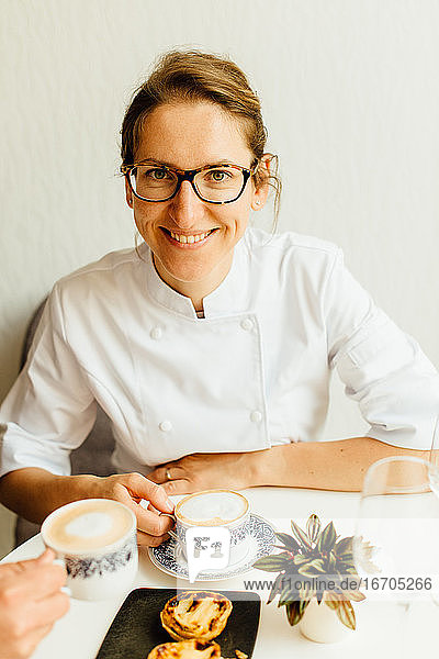 Woman in white uniform smiling while sitting at table with cappuccino