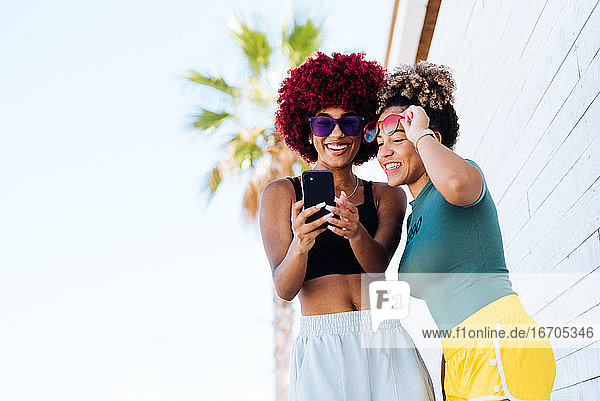 Zwei attraktive Latino-Mädchen mit Afro-Haar suchen Fotos auf Handy.