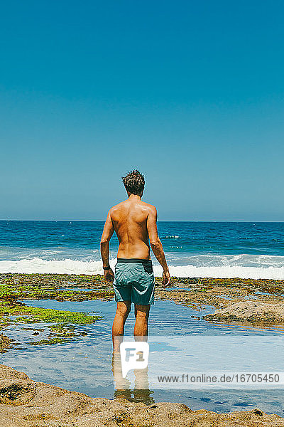 Junger Mann steht im Wasser und schaut von einer Steilküste in Baja auf den Ozean hinaus.